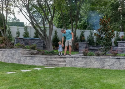A man helps a small child walk down steps in a beautifully landscaped backyard in MD, featuring trees and elegant hardscaping with stone walls.