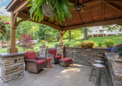 Covered patio with stone walls, red cushioned chairs, lush plants, and a garden view in a sunny backyard featuring expert landscaping from Solid Brick Contracting.