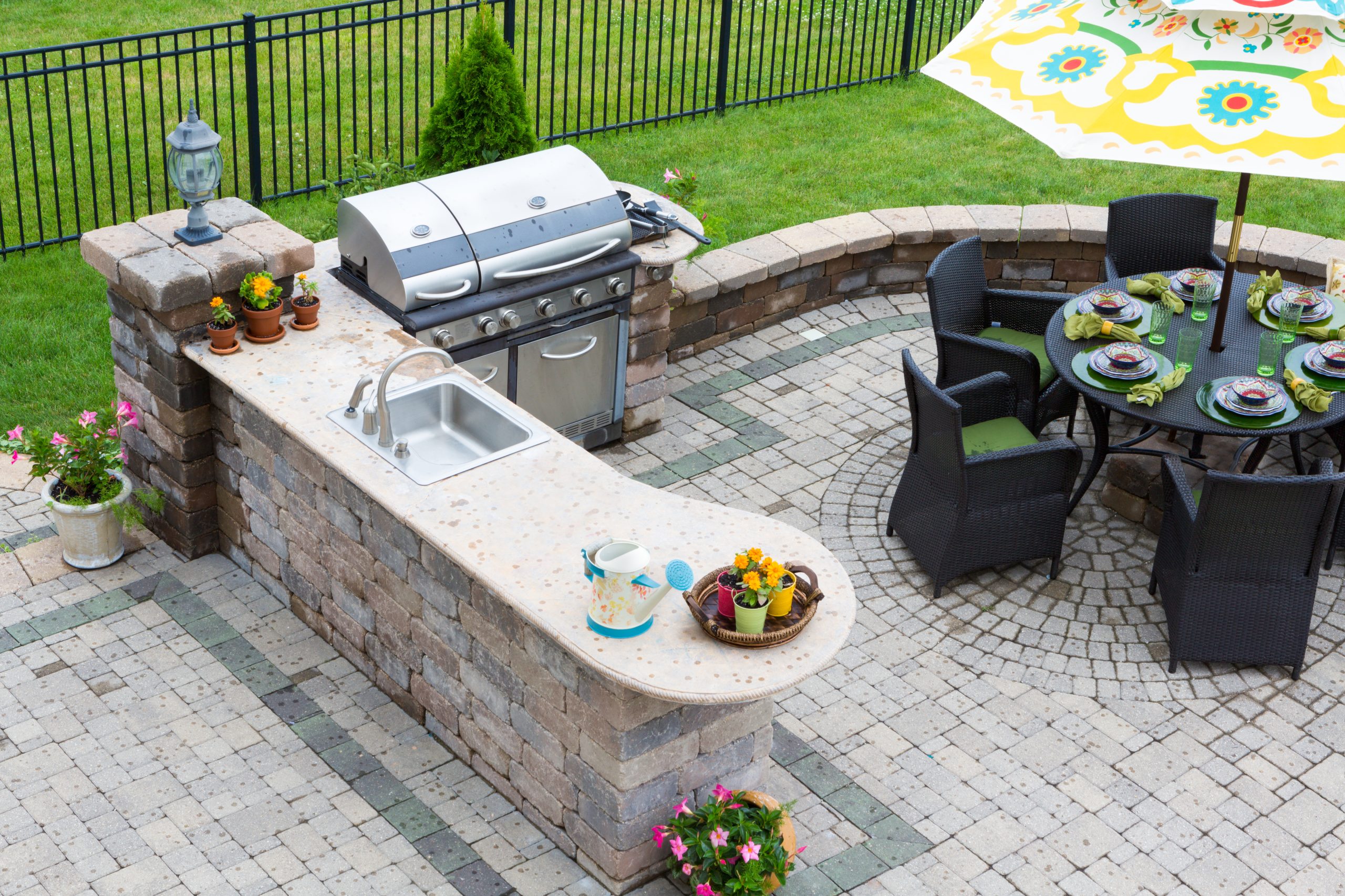 hardscaping contractor: Why Outdoor Kitchens Are the Ultimate Backyard Upgrade for Entertaining Outdoor patio with a grill, sink, and bar area. Dining table with chairs and umbrella. Potted plants and flowers.