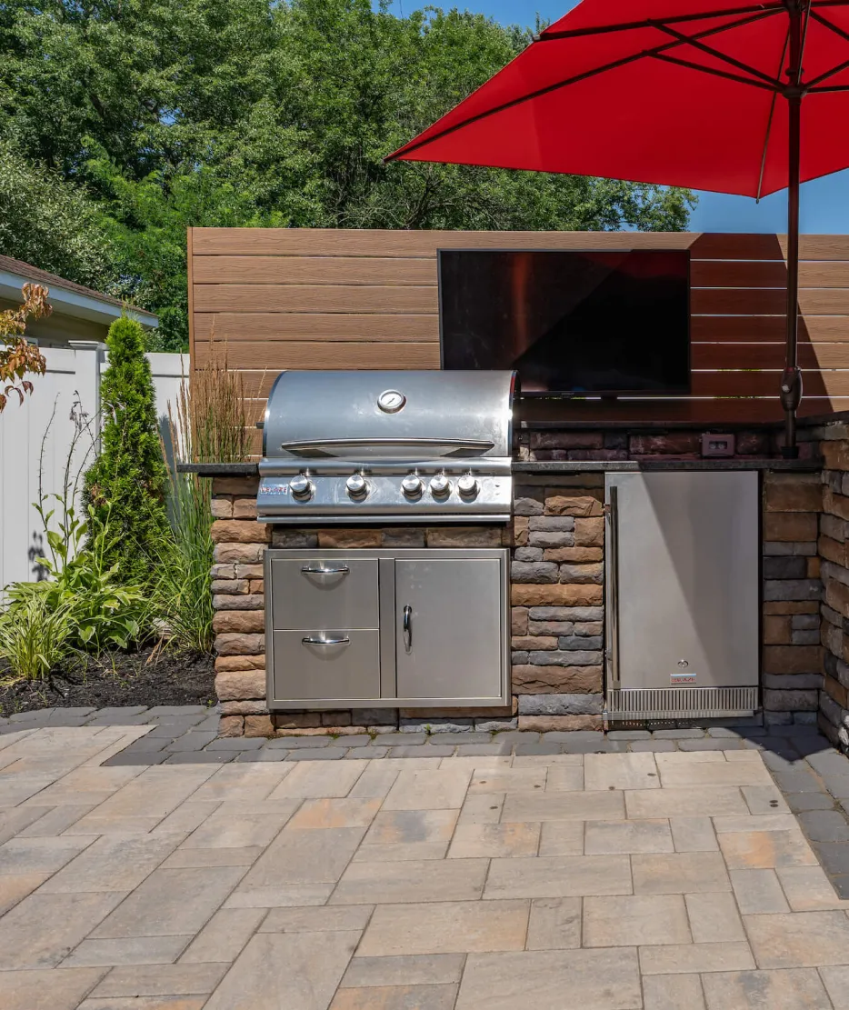 Outdoor kitchen with a stainless steel grill, mini fridge, TV, and a red umbrella on a stone patio.