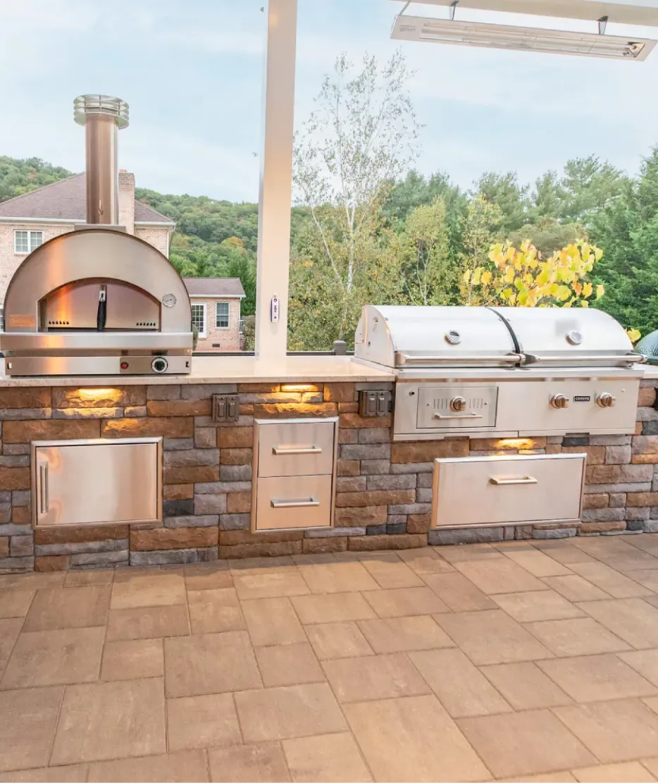 Outdoor kitchen with stone wall, stainless steel appliances, pizza oven, and grill on a tiled patio.