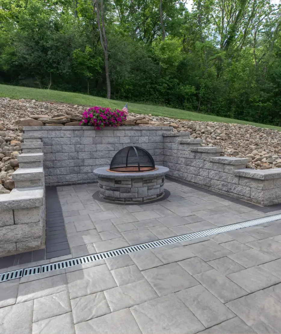 walls-1 Stone patio with a round fire pit, retaining wall, and pink flowers, bordered by rocks and trees.