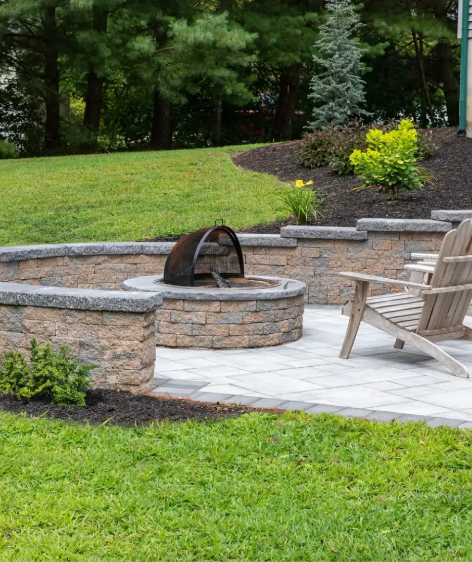 walls-2 Stone patio with a built-in fire pit, wooden chairs, and landscaped greenery in a backyard setting.